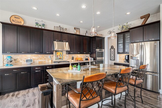7692 Weaver Cir, Castle Rock, CO 80104 - kitchen featuring appliances with stainless steel finishes, under cabinet range hood, light wood-style floors, decorative backsplash, and a kitchen bar