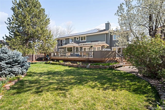 rear view of house with roof mounted solar panels, a chimney, fence, a lawn, and a deck