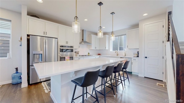 kitchen with appliances with stainless steel finishes, wall chimney exhaust hood, pendant lighting, a kitchen island, and white cabinetry