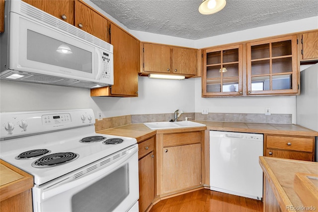 kitchen featuring a sink, a textured ceiling, wood finished floors, white appliances, and glass insert cabinets