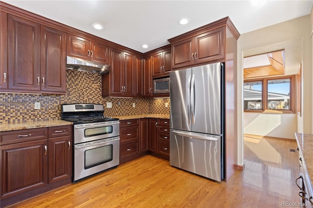 kitchen with under cabinet range hood, light wood finished floors, appliances with stainless steel finishes, and tasteful backsplash