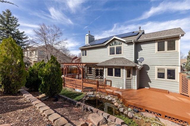 back of house featuring a wooden deck, roof mounted solar panels, a chimney, and a pergola