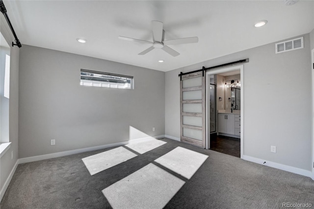 unfurnished bedroom with dark carpet, a barn door, ensuite bath, and ceiling fan