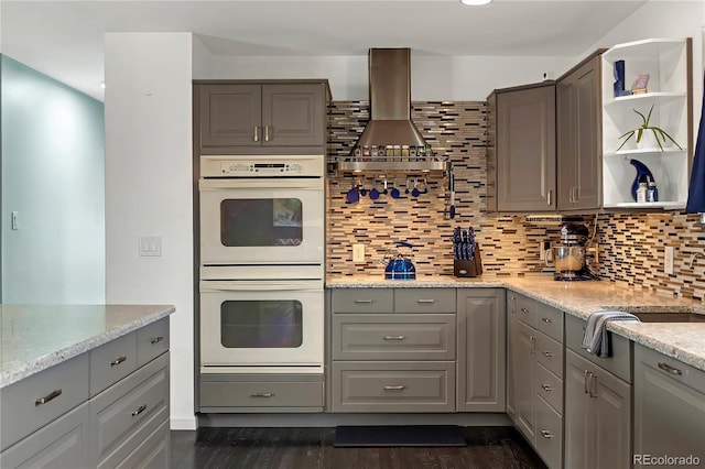 kitchen featuring light stone countertops, double oven, gray cabinets, decorative backsplash, and exhaust hood