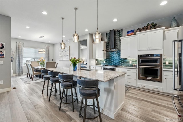 kitchen featuring wall chimney exhaust hood, white cabinets, a sink, multiple ovens, and light stone countertops