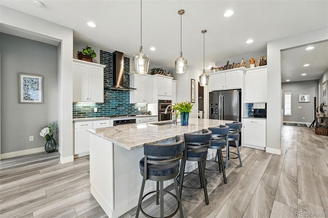 kitchen with stainless steel appliances, wall chimney range hood, light wood-style floors, a kitchen island with sink, and tasteful backsplash