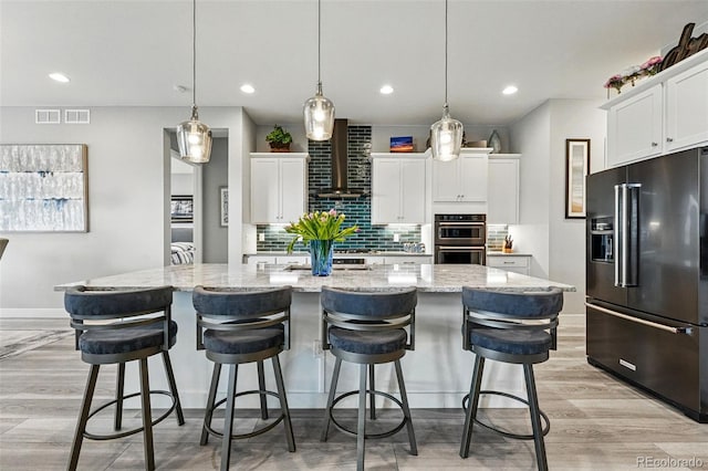 kitchen with wall chimney range hood, multiple ovens, decorative backsplash, high quality fridge, and a center island with sink