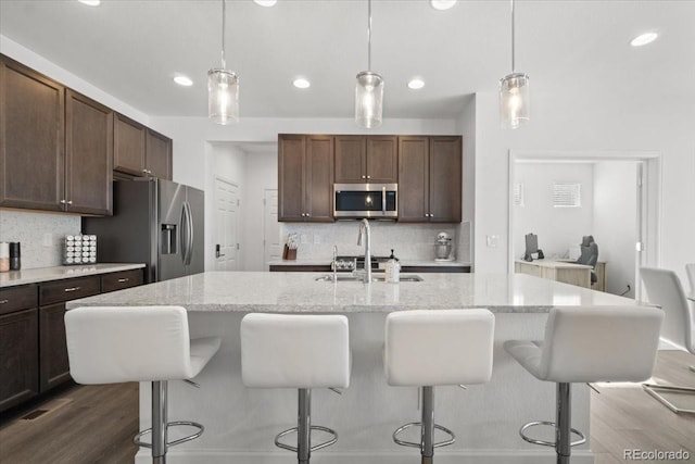 kitchen with appliances with stainless steel finishes, hanging light fixtures, dark brown cabinetry, light stone counters, and an island with sink