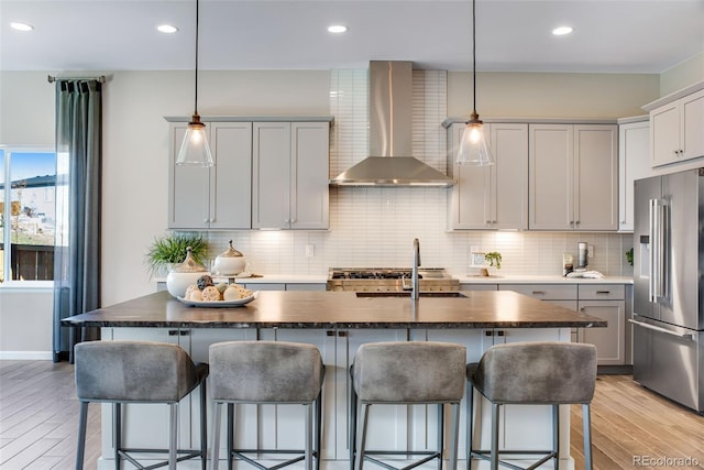 kitchen featuring high end fridge, light hardwood / wood-style floors, a center island with sink, wall chimney exhaust hood, and pendant lighting