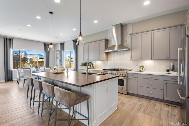 kitchen with stainless steel appliances, wall chimney exhaust hood, sink, an island with sink, and decorative light fixtures