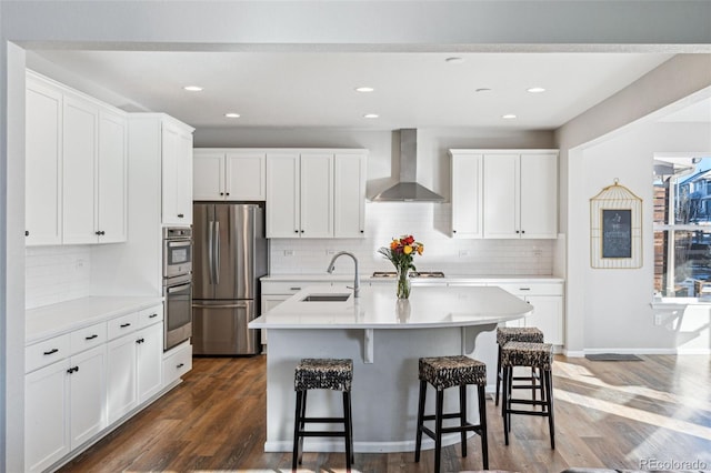 kitchen with wall chimney exhaust hood, sink, a center island with sink, stainless steel appliances, and white cabinets