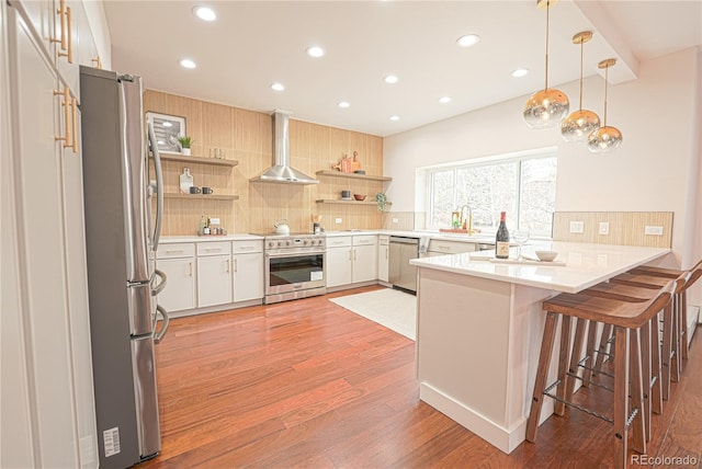 kitchen featuring a kitchen breakfast bar, stainless steel appliances, wall chimney range hood, white cabinetry, and kitchen peninsula