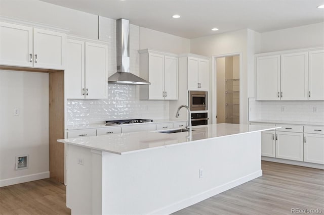 kitchen featuring wall chimney range hood, sink, appliances with stainless steel finishes, a kitchen island with sink, and white cabinets