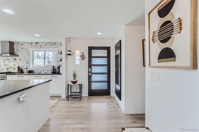 kitchen featuring wall chimney range hood, hanging light fixtures, decorative backsplash, light hardwood / wood-style floors, and white cabinetry