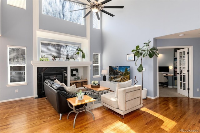 living area with baseboards, a towering ceiling, wood finished floors, a glass covered fireplace, and a ceiling fan