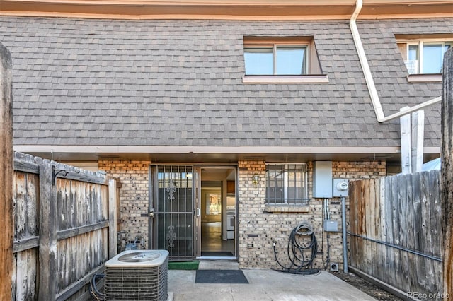 view of exterior entry with brick siding, roof with shingles, central AC, and fence