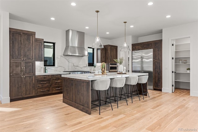 kitchen featuring light wood-type flooring, hanging light fixtures, wall chimney exhaust hood, a center island with sink, and appliances with stainless steel finishes