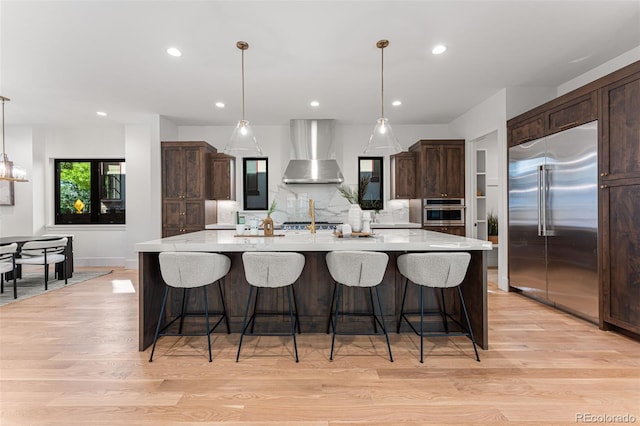 kitchen featuring a large island with sink, hanging light fixtures, wall chimney range hood, and built in fridge