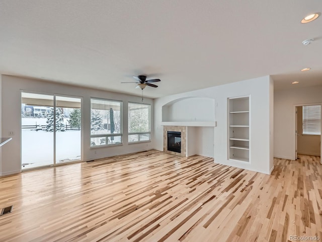 unfurnished living room featuring built in shelves, a tiled fireplace, ceiling fan, and light hardwood / wood-style floors