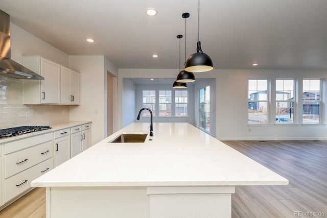 kitchen with gas stovetop, a kitchen island with sink, a sink, decorative backsplash, and wall chimney exhaust hood