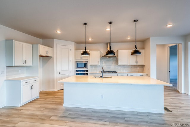 kitchen with a kitchen island with sink, built in microwave, a sink, stainless steel oven, and wall chimney range hood