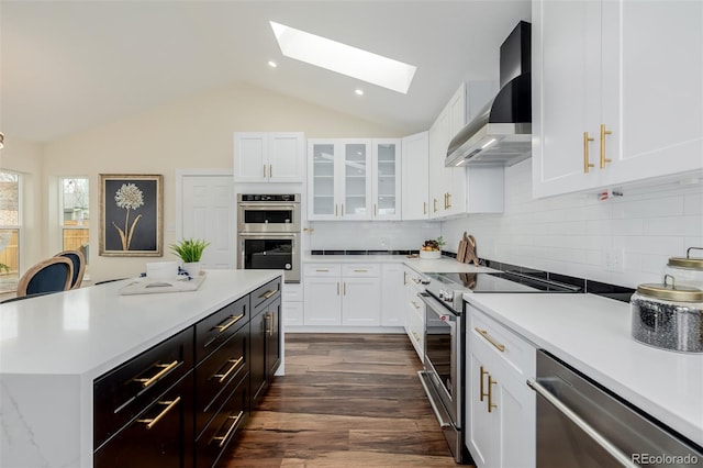 kitchen with dark wood-type flooring, appliances with stainless steel finishes, lofted ceiling with skylight, white cabinets, and wall chimney exhaust hood