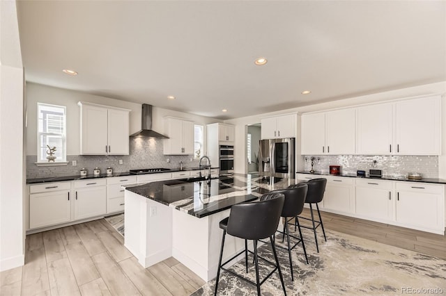 kitchen featuring a breakfast bar, stainless steel appliances, wall chimney range hood, a center island with sink, and white cabinets