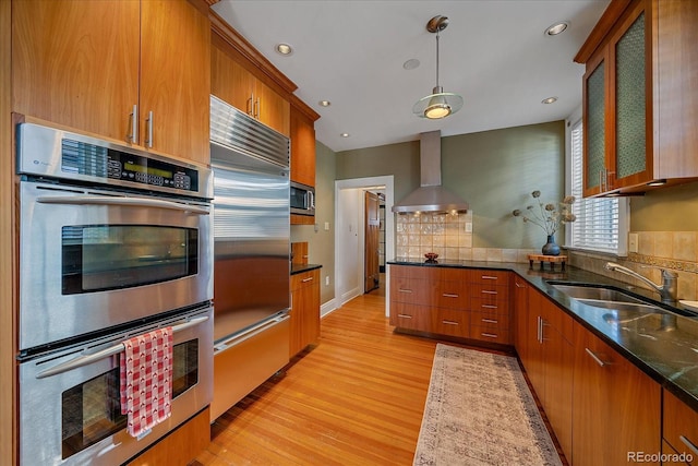 kitchen with wall chimney exhaust hood, sink, light hardwood / wood-style flooring, appliances with stainless steel finishes, and pendant lighting