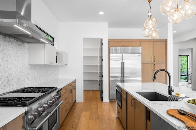 kitchen with wall chimney exhaust hood, built in appliances, a sink, a chandelier, and light stone countertops