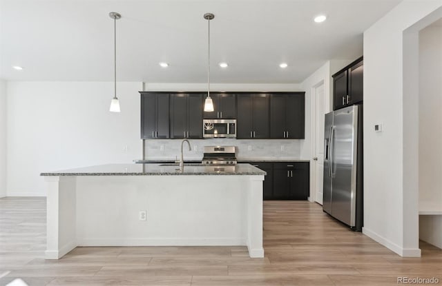 kitchen with light stone counters, an island with sink, appliances with stainless steel finishes, and pendant lighting