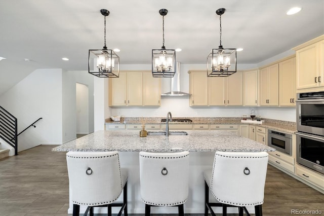 6692 S Robertsdale Way, Aurora, CO 80016 - kitchen featuring cream cabinetry, wall chimney range hood, stainless steel appliances, a chandelier, and an island with sink