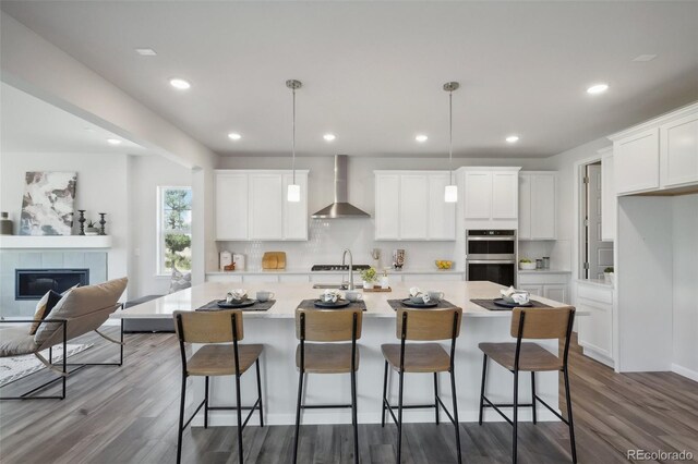 kitchen with white cabinets, wall chimney exhaust hood, an island with sink, and decorative light fixtures