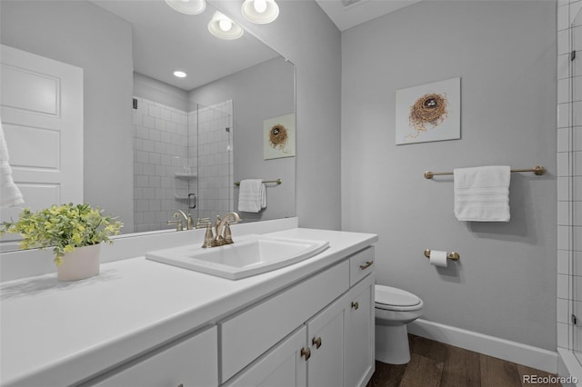 bathroom with toilet, wood finished floors, vanity, baseboards, and a tile shower