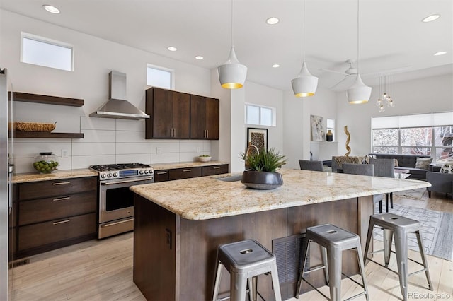 kitchen featuring stainless steel gas range, wall chimney exhaust hood, dark brown cabinetry, open shelves, and a kitchen breakfast bar