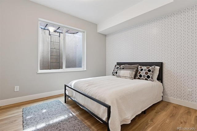 bedroom featuring baseboards, wallpapered walls, an accent wall, and wood finished floors