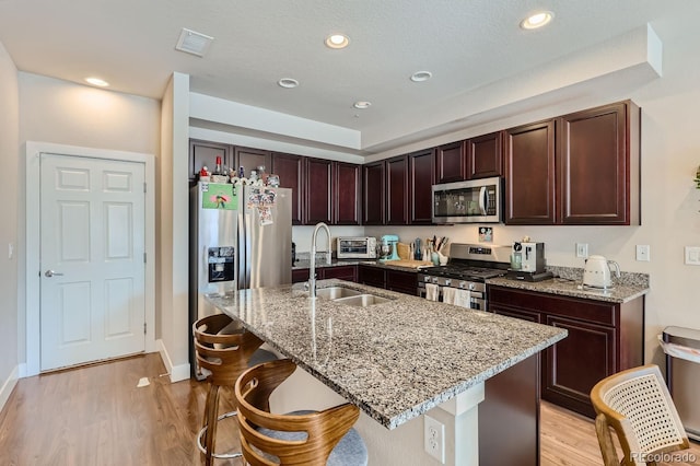 kitchen featuring light stone counters, a kitchen bar, a kitchen island with sink, appliances with stainless steel finishes, and light wood-type flooring