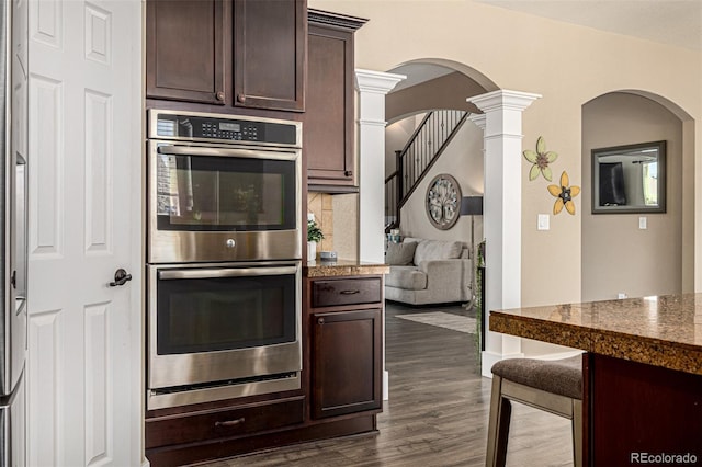 kitchen featuring double oven, decorative columns, dark brown cabinets, dark wood-style floors, and arched walkways