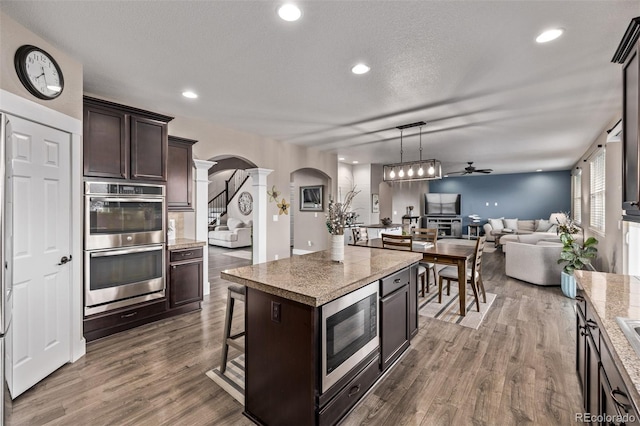 kitchen featuring decorative columns, arched walkways, appliances with stainless steel finishes, dark brown cabinetry, and ceiling fan