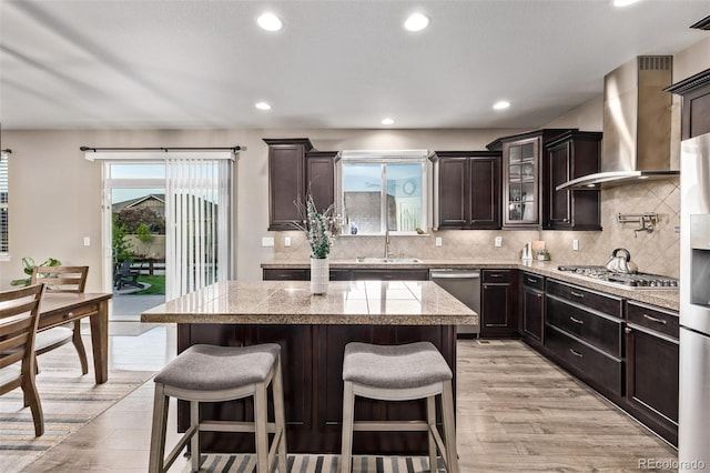 kitchen featuring wall chimney exhaust hood, stainless steel appliances, light wood finished floors, a sink, and a kitchen bar