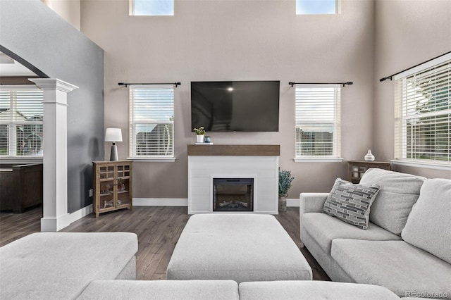 living area featuring a high ceiling, wood finished floors, ornate columns, baseboards, and a glass covered fireplace