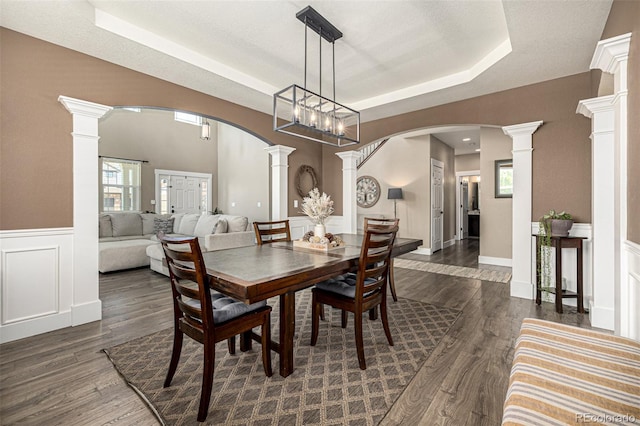dining room with ornate columns, arched walkways, a raised ceiling, dark wood-style flooring, and a wainscoted wall