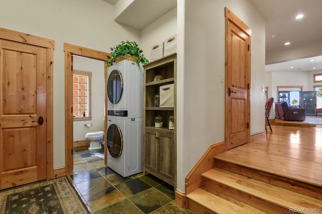 clothes washing area with recessed lighting, stone tile flooring, stacked washer / dryer, laundry area, and baseboards