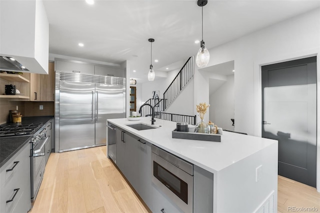 kitchen featuring sink, built in appliances, an island with sink, decorative light fixtures, and light wood-type flooring