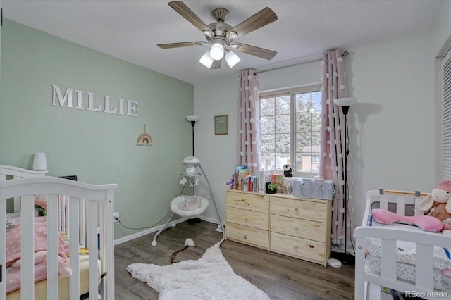 bedroom with a nursery area, a ceiling fan, baseboards, and dark wood-style flooring