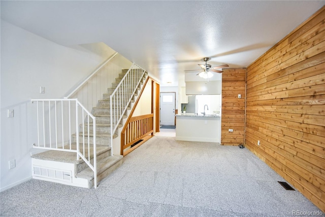 unfurnished living room with light colored carpet, visible vents, ceiling fan, wood walls, and stairs