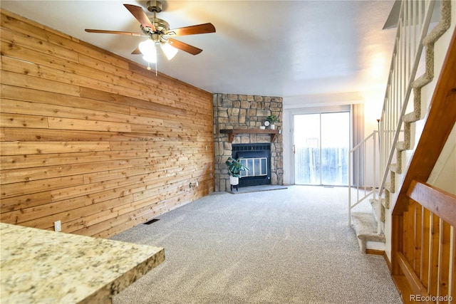 unfurnished living room with ceiling fan, carpet floors, wood walls, a fireplace, and stairs