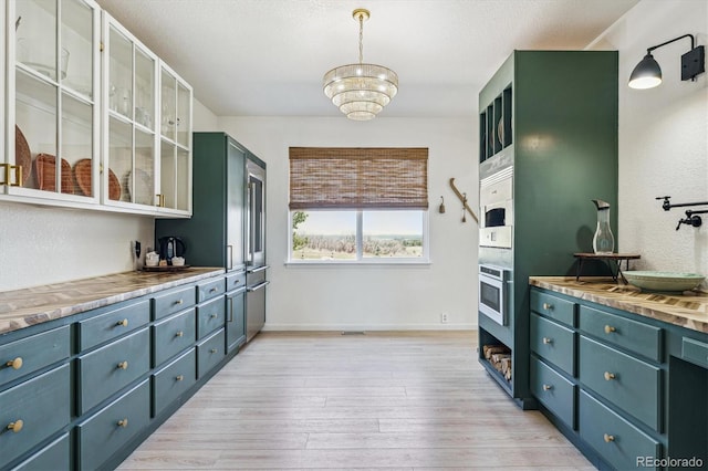 kitchen with light wood finished floors, a chandelier, glass insert cabinets, baseboards, and wall oven