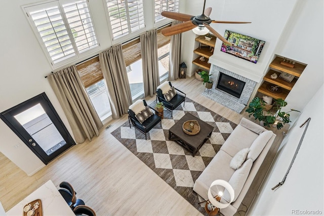 living area featuring wood finished floors, a ceiling fan, a high ceiling, visible vents, and a glass covered fireplace