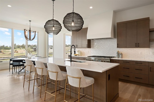 kitchen featuring light wood-type flooring, tasteful backsplash, custom range hood, sink, and a center island with sink