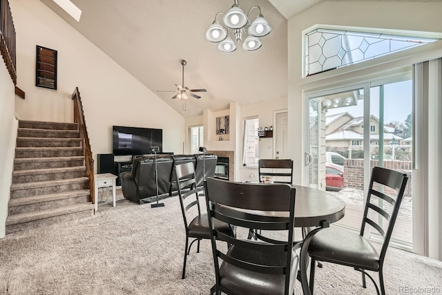 carpeted dining room featuring ceiling fan with notable chandelier, high vaulted ceiling, stairway, and a tile fireplace